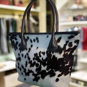 May include: A white and black cowhide tote bag with black leather handles. The bag has a large, open top and a spacious interior.