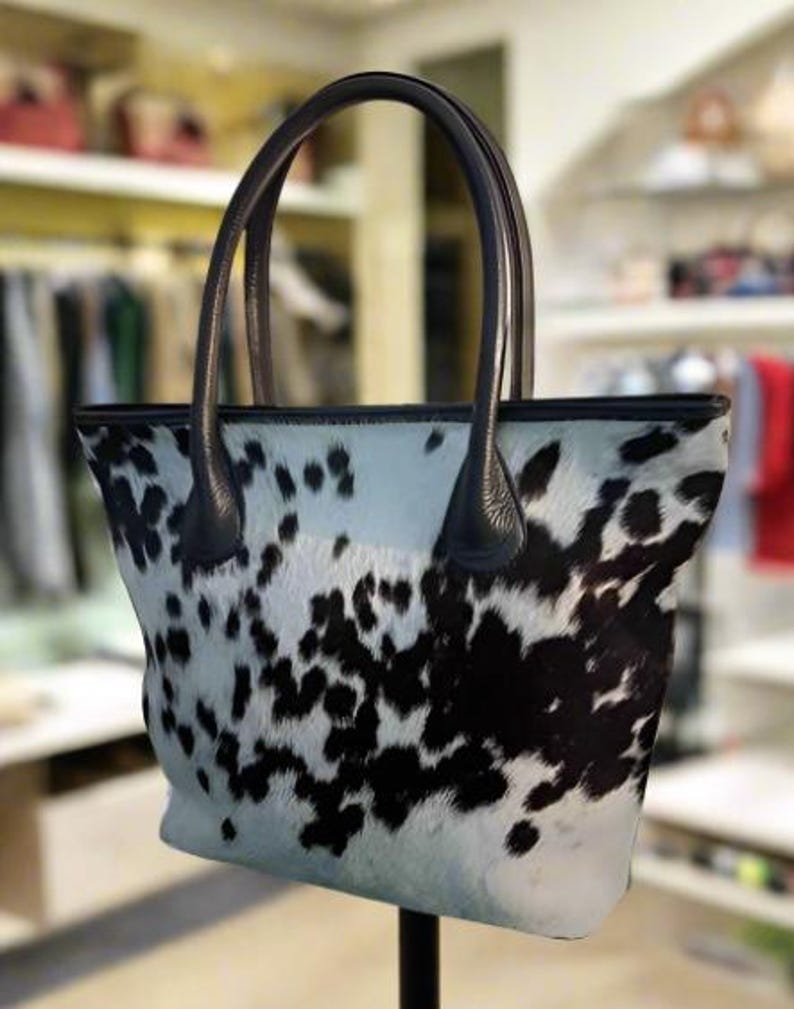 May include: A white and black cowhide tote bag with black leather handles. The bag has a large, open top and a spacious interior.