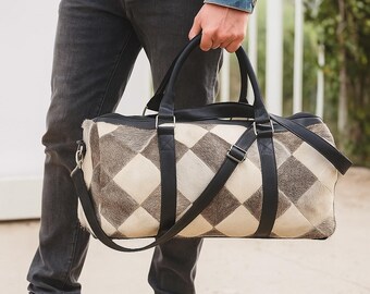 Könnte beinhalten: Eine Reisetasche mit einem geometrischen Muster aus abwechselnd schwarzen und cremefarbenen Rauten. Die Tasche hat schwarze Ledergriffe und einen Schultergurt. Die Tasche wird von einer Person getragen, die Jeans und eine Jeansjacke trägt.