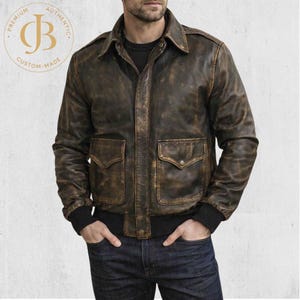 May include: A distressed brown leather jacket with two front pockets and a collared neckline. The jacket has a ribbed hem and cuffs. The image includes a circular logo with the text "PREMIUM AUTHENTIC CUSTOM-MADE" and the letters "JB".