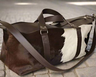 Könnte beinhalten: Eine braune Ledertasche mit einem weißen und braunen Kuhfellmuster. Die Tasche hat einen langen Schultergurt und zwei Tragegriffe oben.
