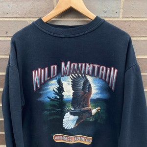 Może przedstawiać: Czarna bluza z długim rękawem z nadrukiem graficznym orła lecącego nad g&oacute;rskim krajobrazem. Tekst "WILD MOUNTAIN" jest nadrukowany w czerwono-białym łuku nad orłem. Tekst "WILDERNESS EXPEDITION" jest nadrukowany w czerwono-białym prostokącie pod orłem.