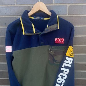 Peut inclure: Un pull en polaire bleu et vert avec une bordure jaune. Le pull a un col boutonné et un patch sur la poitrine qui dit "Polo Sport". La manche droite a un graphisme blanc qui dit "RLPC67" et un patch plus petit qui dit "Polo Ralph Lauren Established 1967".