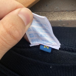 Może przedstawiać: Czarny T-shirt z niebieską metką pielęgnacyjną, na kt&oacute;rej widnieje napis "Prać w pralce w zimnej wodzie, suszyć w suszarce bębnowej w niskiej temperaturze, nie wybielać, nie prasować, nie czyścić chemicznie" oraz niebieską metką rozmiaru z napisem "M".