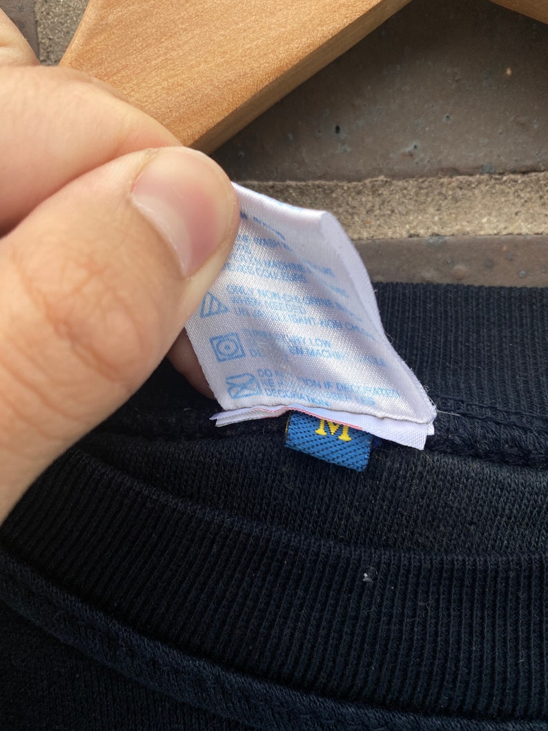 Może przedstawiać: Czarny T-shirt z niebieską metką pielęgnacyjną, na kt&oacute;rej widnieje napis "Prać w pralce w zimnej wodzie, suszyć w suszarce bębnowej w niskiej temperaturze, nie wybielać, nie prasować, nie czyścić chemicznie" oraz niebieską metką rozmiaru z napisem "M".