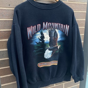 Może przedstawiać: Czarna bluza z nadrukiem graficznym orła lecącego nad sceną leśną. Tekst "WILD MOUNTAIN" jest nadrukowany białym, srebrnym i czerwonym kolorem. Tekst "WILDERNESS EXPEDITION" jest nadrukowany brązowym kolorem na czerwonej wstążce.