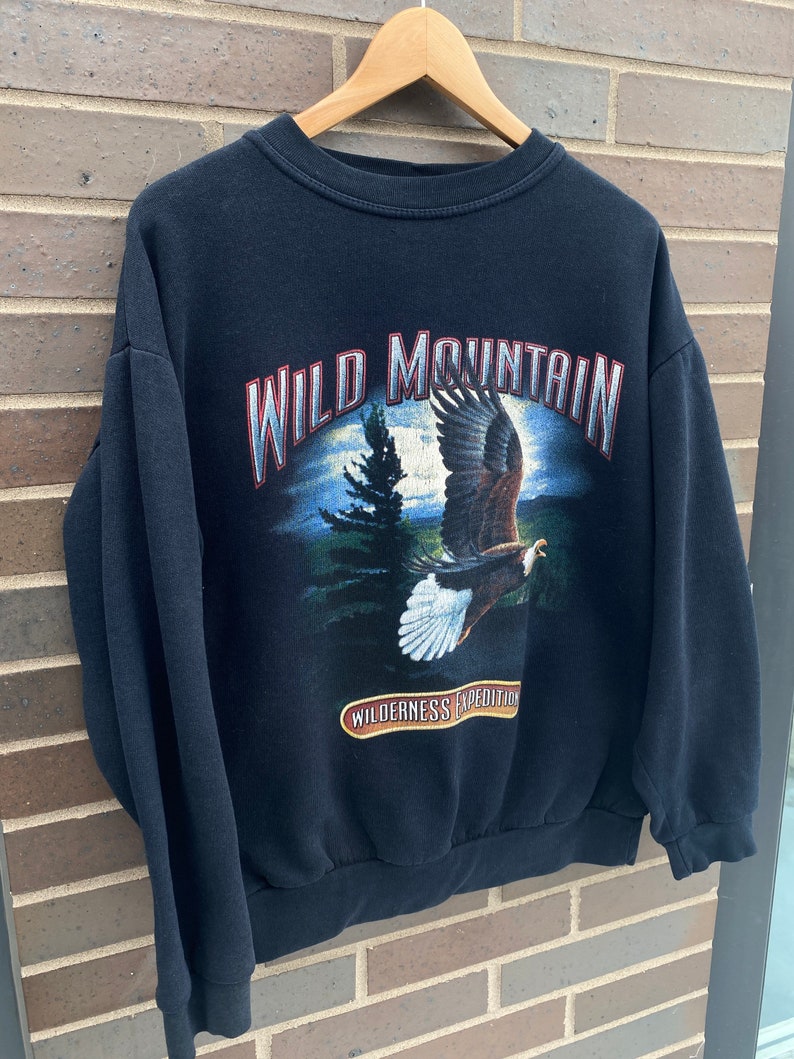 Może przedstawiać: Czarna bluza z nadrukiem graficznym orła lecącego nad sceną leśną. Tekst "WILD MOUNTAIN" jest nadrukowany białym, srebrnym i czerwonym kolorem. Tekst "WILDERNESS EXPEDITION" jest nadrukowany brązowym kolorem na czerwonej wstążce.