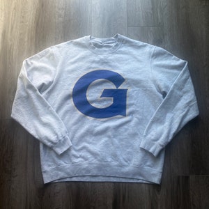 Peut inclure: Un sweat-shirt gris avec un logo "G" bleu sur le devant. Le logo est bordé d'or et comporte un symbole de marque déposée.
