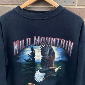 Może przedstawiać: Czarny sweter z nadrukiem graficznym przedstawiającym orła bielika lecącego nad pasmem g&oacute;rskim. Tekst "WILD MOUNTAIN" jest nadrukowany na czerwono i biało nad orłem. Tekst "WILDERNESS EXPEDITION" jest nadrukowany na czerwono i biało pod orłem.
