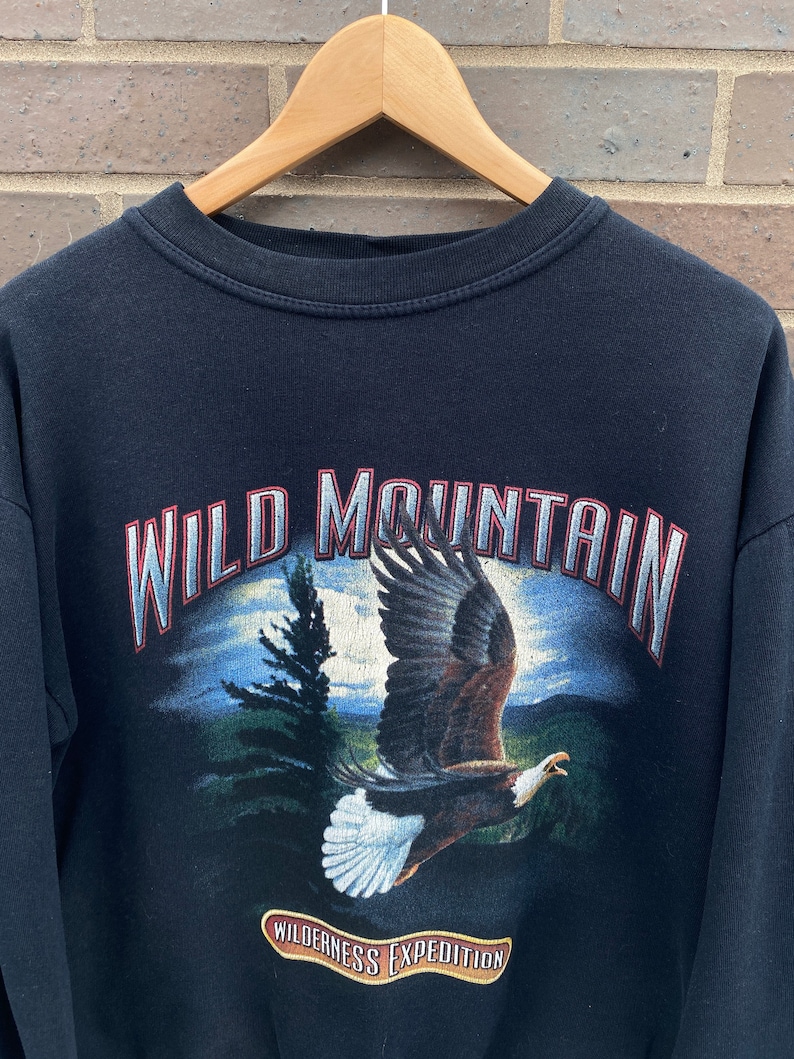 Może przedstawiać: Czarny sweter z nadrukiem graficznym przedstawiającym orła bielika lecącego nad pasmem g&oacute;rskim. Tekst "WILD MOUNTAIN" jest nadrukowany na czerwono i biało nad orłem. Tekst "WILDERNESS EXPEDITION" jest nadrukowany na czerwono i biało pod orłem.