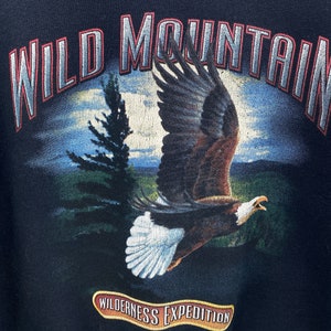 Może przedstawiać: Czarna bluza z nadrukiem graficznym orła lecącego nad pasmem g&oacute;rskim. Tekst "WILD MOUNTAIN" jest nadrukowany na czerwono i biało nad orłem. Tekst "WILDERNESS EXPEDITION" jest nadrukowany na złoto pod orłem.