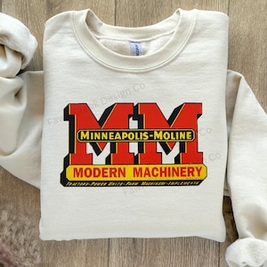 May include: Beige sweatshirt with a red and yellow logo that says "Minneapolis-Moline" and "Modern Machinery" with the text "Tractors-Power Units-Farm Machinery-Implements" underneath.
