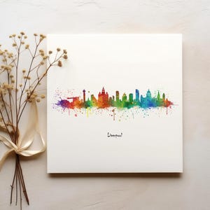 Puede incluir: Una pintura de acuarela del horizonte de Liverpool en colores arcoíris. La pintura está en una tarjeta blanca con la palabra "Liverpool!" escrita debajo del horizonte.