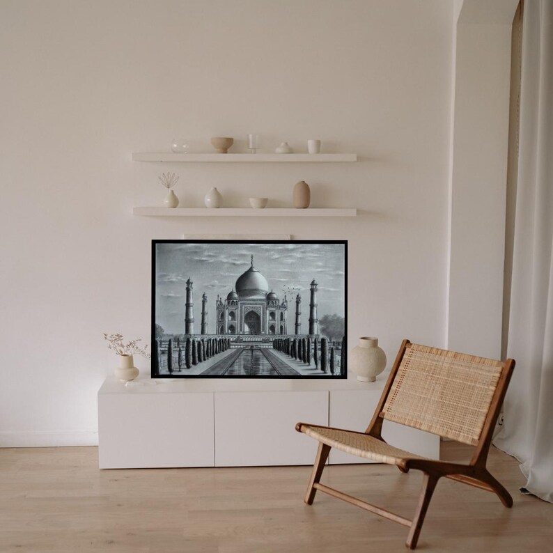 May include: A black and white drawing of the Taj Mahal, framed and hanging on a white wall in a modern living room. The drawing is in a black frame and is centred on the wall. There is a white shelf above the drawing with decorative objects on it. A wicker chair is in the foreground.