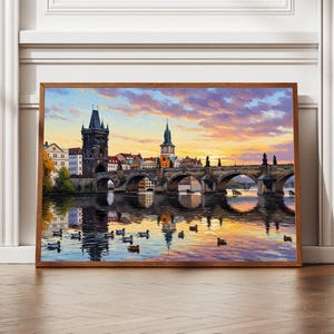 Pode incluir: Uma pintura emoldurada retrata uma ponte de pedra pitoresca sobre um rio ao pôr do sol. A obra apresenta uma torre escura, edifícios e um céu colorido com reflexos na água. Os patos nadam no rio.