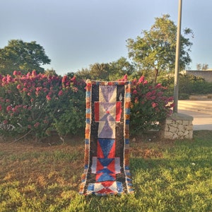 Puede incluir: Una alfombra colorida tejida a mano con patrones geométricos en tonos rojos, azules, naranjas y blancos. La alfombra está colocada en un césped verde frente a un arbusto con flores rosas.
