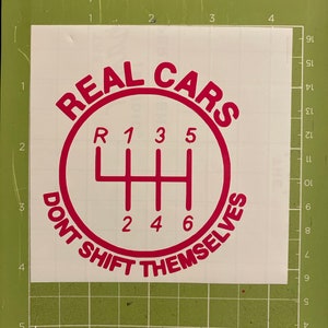 May include: A red vinyl decal with the text "REAL CARS DON'T SHIFT THEMSELVES" in a circle around a stylized gear shift pattern with the numbers 1, 2, 3, 4, 5, and 6.