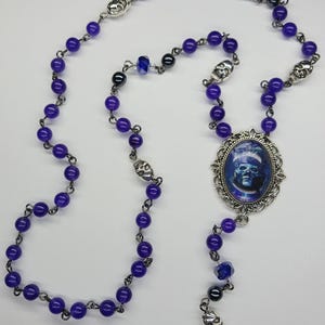 May include: A purple and silver rosary necklace with a skull charm and a silver pendant featuring a blue and white image of a skull.