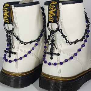 Puede incluir: Un par de botas de cuero blanco con suelas y cordones negros. Las botas están adornadas con cadenas de metal negro, alambre de púas y cadenas de cuentas moradas estilo rosario. Cada bota tiene un colgante de cruz de metal negro.