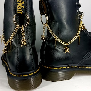 Puede incluir: Un par de botas de cuero negro con cadenas de metal dorado sujetas a las trabillas del talón. Las cadenas están adornadas con pequeñas púas doradas y dijes en forma de estrella, creando un acento metálico decorativo en el calzado.