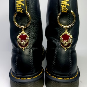 Puede incluir: Un par de botas de cuero negro con costuras amarillas y una trabilla amarilla en el talón con el texto AirWair. Cada bota está adornada con un anillo de metal dorado que sostiene un colgante ovalado con esmalte rojo y un motivo de murciélago.