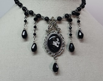 May include: A black choker necklace with a silver pendant featuring a cameo with a black and white silhouette design. The pendant is surrounded by a decorative silver frame and hangs from a chain with black beads. The necklace has multiple black bead drops.