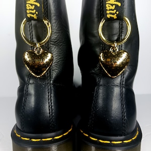 May include: A pair of black leather boots featuring yellow stitching along the soles and black heel loops with yellow text. Each boot is adorned with a gold-toned metal ring attached to the heel loop, holding a metallic heart-shaped charm.