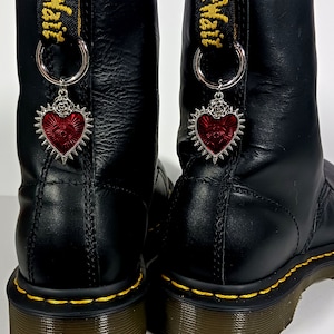Może przedstawiać: Para czarnych skórzanych butów z czerwonymi zawieszkami w kształcie serca z wzorem oka. Buty mają żółte szwy i sznurowadła.
