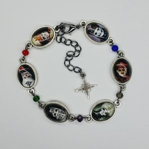 Peut inclure: Un bracelet à breloques argenté avec un charme étoile et des breloques ovales représentant des images en noir et blanc de personnes. Le bracelet est orné de perles rouges, bleues et vertes.