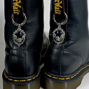 Pode incluir: Botas de couro preto com costuras amarelas e um amuleto de morcego prateado preso a cada bota.