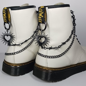 Peut inclure: Deux bottes en cuir blanc avec des semelles noires et des tirettes jaunes et noires. Les bottes sont ornées de chaînes en métal noir, de fil de fer barbelé et de breloques en forme de cœur.