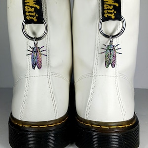 May include: A pair of white leather boots featuring black heel pull tabs with yellow text. Each boot is adorned with a silver-toned metal ring attached to the pull tab, from which hangs an iridescent, multi-coloured cicada-shaped metal charm.