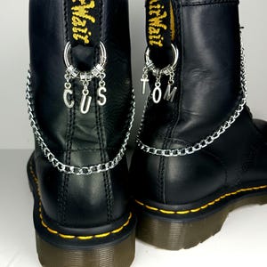 Puede incluir: Botas de cuero negro con cadenas y dijes plateados. Los dijes deletrean "TOM" y "CUS".