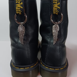 Może przedstawiać: Para czarnych skórzanych butów z żółtymi szwami i uchwytami. Każdy but ma srebrny urok skrzydła wiszący z metalowego pierścienia przymocowanego do uchwytu. Tło jest gładkie białe.