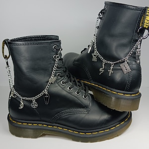 Könnte beinhalten: Schwarze Lederstiefel mit silbernen Ketten und Anhängern, darunter ein Halbmond, ein Pentagramm, ein Sarg und eine Kassette. Die Stiefel haben gelbe Nähte und eine schwarze Gummisohle.