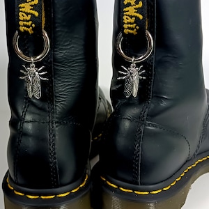 Może przedstawiać: Czarne skórzane buty z żółtymi szwami i srebrnymi zawieszkami w kształcie owadów przywiązanymi do sznurowadeł. Buty mają grubą podeszwę i zaokrąglony nosek.