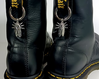 May include: Black leather boots with yellow stitching and silver insect charms attached to the laces. The boots have a thick sole and a rounded toe.