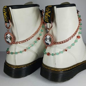 May include: Pair of white leather boots with black soles and yellow stitching. Each boot features a decorative chain with a cat cameo pendant, green beads, and red accents. The boots have black and yellow pull tabs.