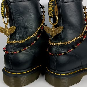 Könnte beinhalten: Schwarze Lederstiefel mit goldenen Ketten- und Perlenakzenten. Die Stiefel haben einen goldenen Schmetterlingsanhänger an jedem Stiefel. Die Stiefel haben ein gelbes AirWair-Etikett an jedem Stiefel.