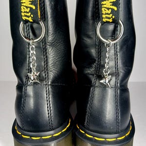 Puede incluir: Botas de cuero negro con costuras amarillas y una presilla amarilla y negra. Cada bota tiene una cadena plateada con un dije en forma de estrella, sujeta al anillo de la presilla.