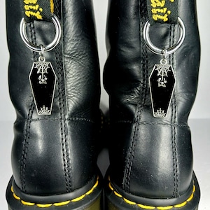 May include: Close-up of black leather boots with yellow stitching and a black coffin-shaped charm hanging from each boot. The charms have a spiderweb design and a spider. The boots have a black and yellow pull tab.