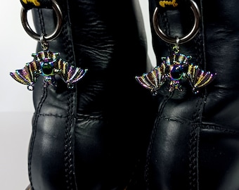 May include: A pair of black leather boots with yellow stitching and a black strap with yellow text. Each boot has a silver charm in the shape of a bat with iridescent rainbow coloring.