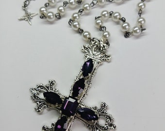 May include: A silver necklace with a large, ornate, upside-down cross pendant. The cross is adorned with purple gemstones. The necklace is made of a silver chain and white pearl beads.