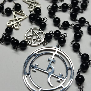 May include: A silver necklace with black beads and silver charms. The charms include a pentagram, a circle with a cross and a crescent moon, and a symbol with a cross and a crescent moon.