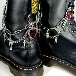 Pode incluir: Botas de couro preto com costuras amarelas, adornadas com correntes prateadas e berloques góticos. Os berloques incluem um esqueleto, cruzes, um coração vermelho, um caixão e um pentagrama.