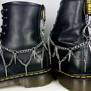 May include: A pair of black leather boots featuring yellow stitching along the soles and yellow heel pull tabs. Each boot is adorned with a silver-toned metal chain harness that drapes across the side, featuring dangling metal spikes.