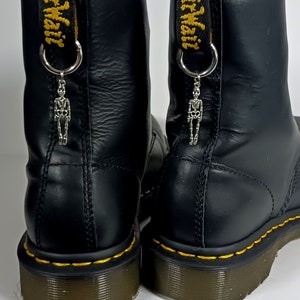 Op de afbeelding: Zwarte leren laarzen met gele stiksels en een skeletbedel bevestigd aan de rits. De rits heeft de tekst "Dr. Martens" geborduurd.