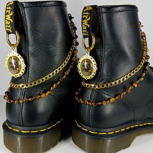Puede incluir: Botas de cuero negro con cadenas doradas y cuentas marrones. Las botas tienen un dije dorado con un diseño de calavera y el texto "Air Wait" en la parte posterior de cada bota.