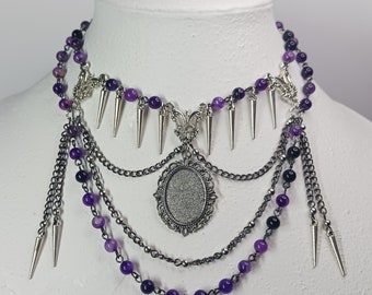 May include: A layered silver necklace with purple beads and a silver pendant with a sparkly center. The necklace has multiple chains and spikes.