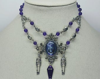 May include: A silver and purple necklace with a cameo featuring a spooky face, two dangling skeletons, and a coffin pendant.